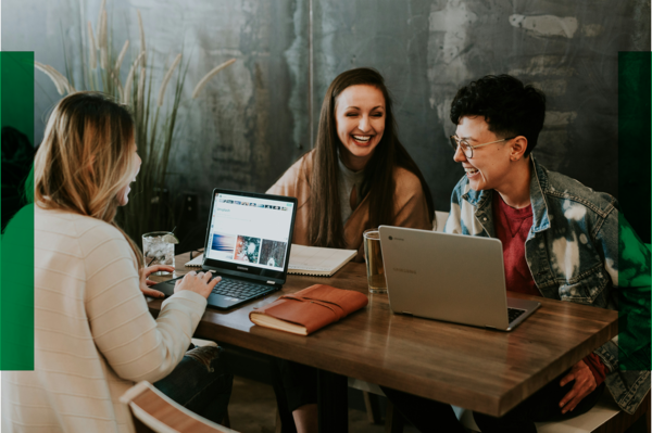 Drei Frauen sitzen zusammen am Tisch und sind miteinander im Gespräch. Vor ihnen stehen Laptops, Bücher und Getränke.