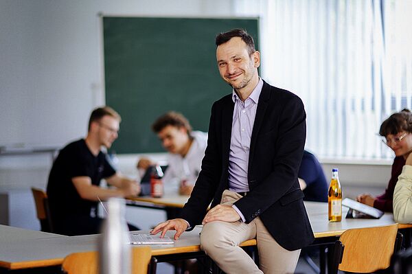 Mann sitzt auf Tisch in Seminarraum, im Hintergrund Studierende